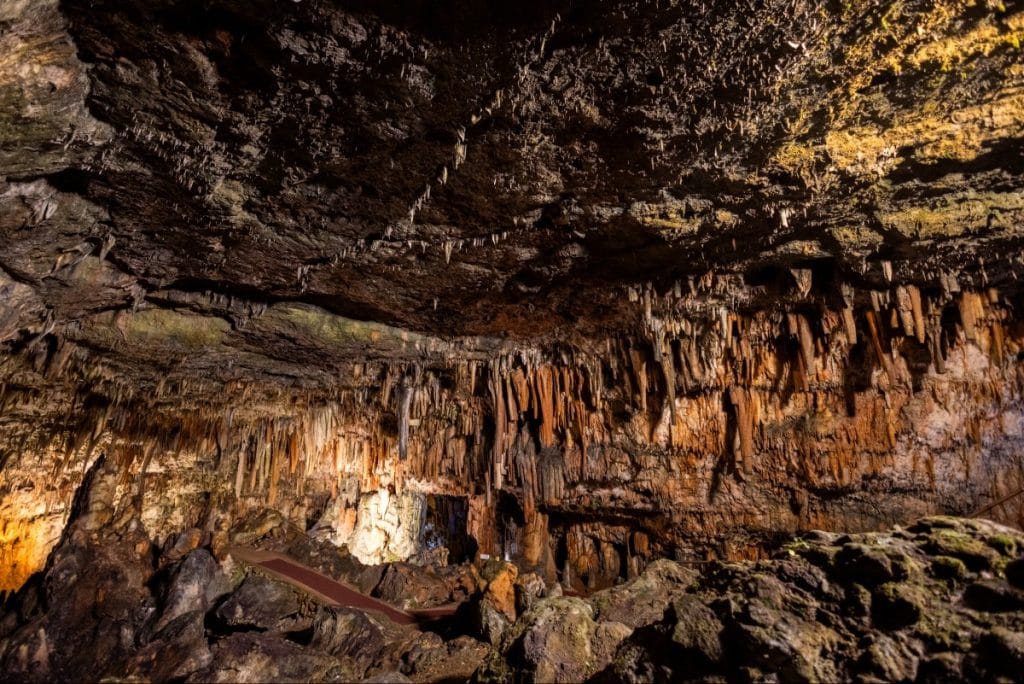 Interior of Drogarati Cave in Kefalonia, Greece, showcasing dramatic stalactites hanging from the ceiling and stalagmites rising from the rocky cave floor. Warm lighting highlights the rich orange and brown tones of the limestone formations, creating a striking underground landscape.