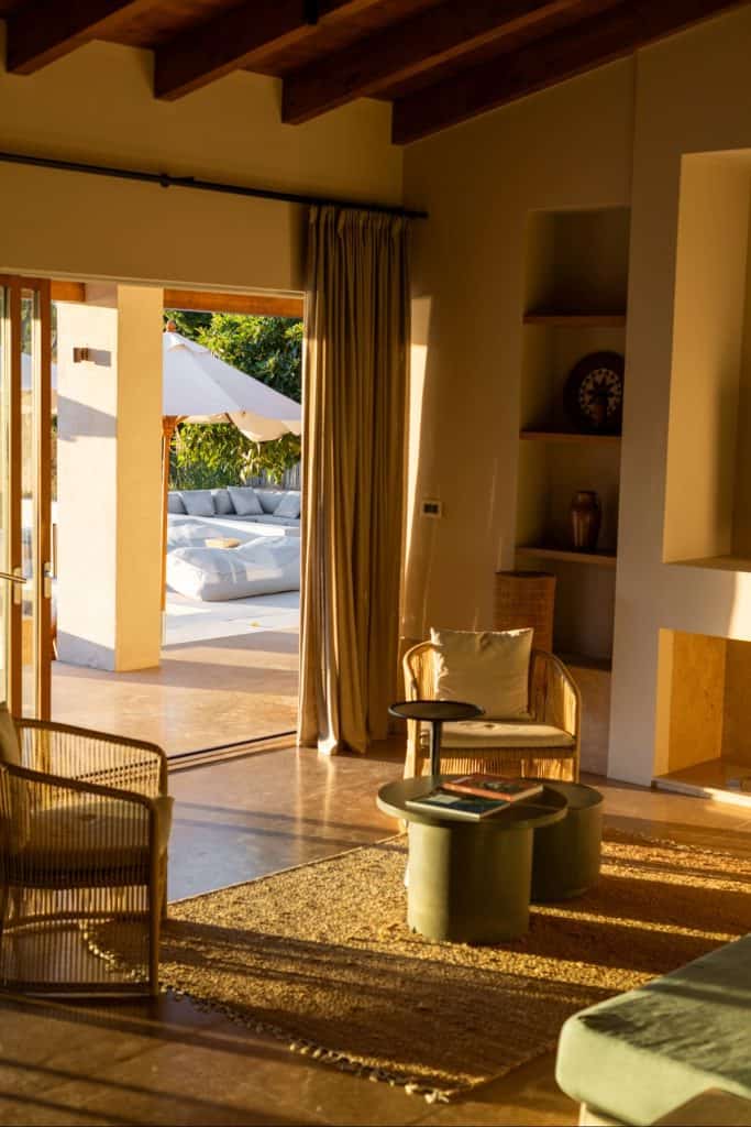Warmly lit interior of a modern Mediterranean villa with natural tones, featuring wicker furniture, a textured rug, and an open sliding door leading to a sun-drenched patio with lounge chairs and umbrellas.