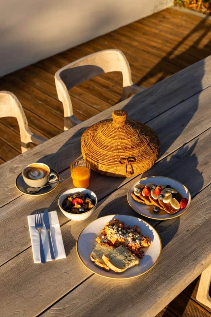 Outdoor breakfast setup on a wooden table with dishes including a cappuccino, fresh juice, yogurt with berries, savory pancakes with cheese, and a plate of mini pancakes topped with fruit. A woven basket food cover adds a rustic touch under the morning sunlight.
