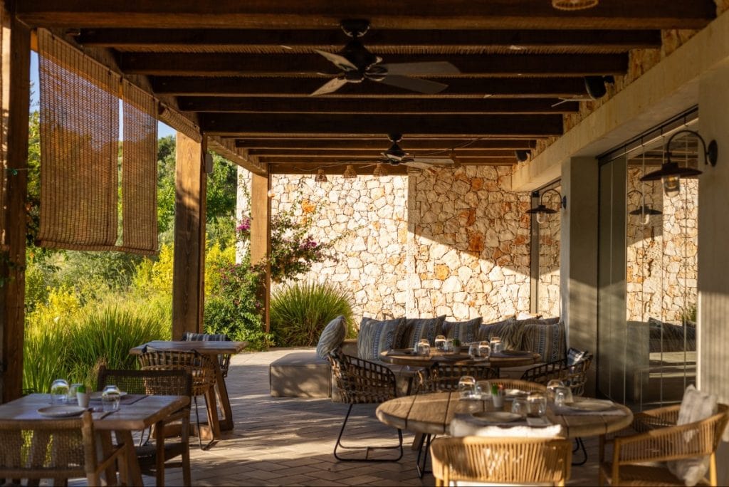Shaded outdoor dining area of a Mediterranean-style villa or restaurant in Kefalonia, Greece, featuring natural wood beams, wicker chairs, and stone walls. Tables are neatly set, and cozy cushioned seating lines the back wall, surrounded by lush garden greenery and soft golden sunlight.
