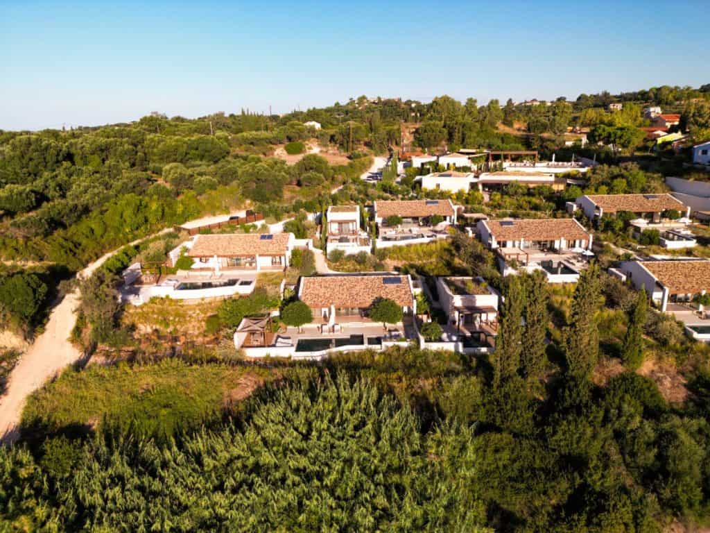 She Knows Alt Text said:

Aerial view of a luxury villa complex nestled in the green hills of Kefalonia, Greece, featuring terracotta-roofed buildings, private pools, and landscaped gardens. The layout is surrounded by dense vegetation and olive trees, blending seamlessly into the natural Mediterranean landscape.