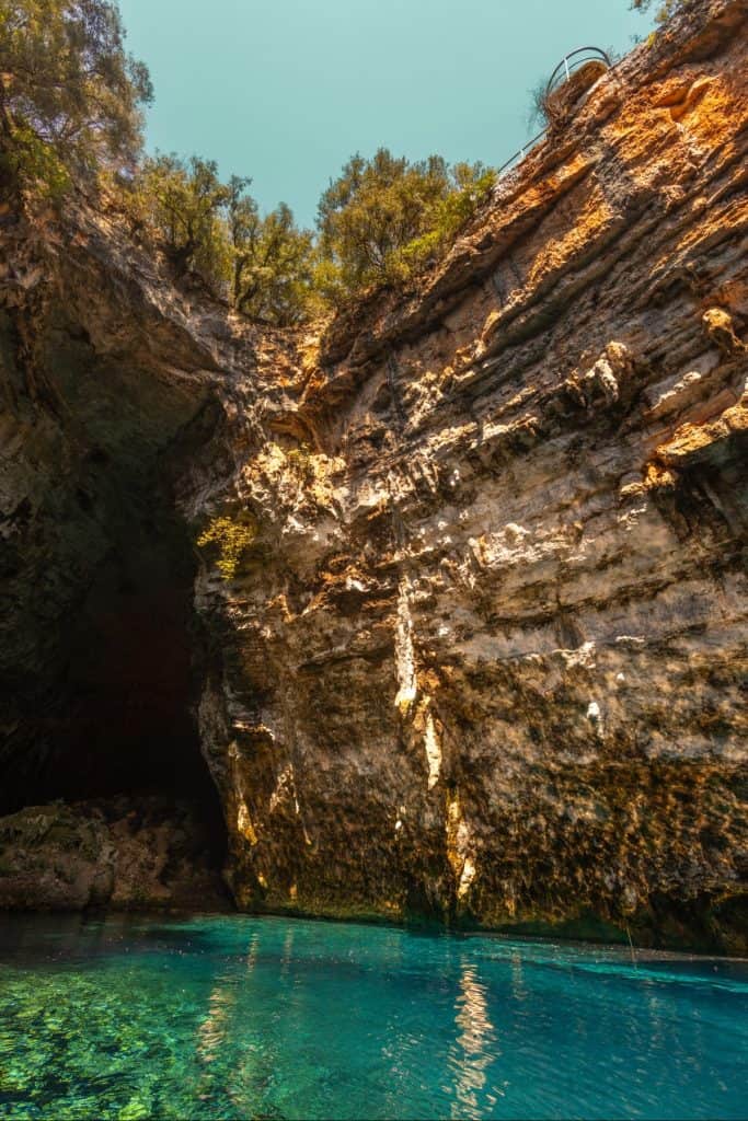 Close-up view of the sunlit limestone walls inside Melissani Cave in Kefalonia, Greece, with turquoise water at the base and trees growing above the cliff edge. The texture of the rock glows in warm tones under the daylight filtering through the cave’s open ceiling.