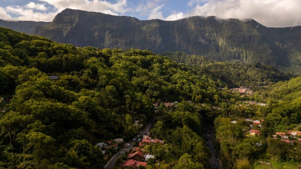 Aerial view of a small village nestled in a deep green valley, surrounded by forested mountains with steep cliffs and scattered clouds.