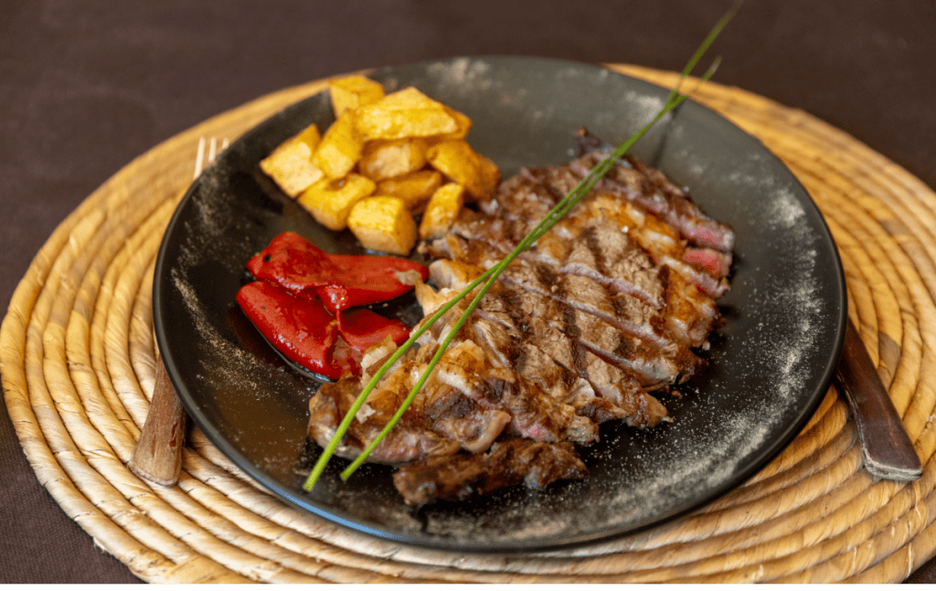 A black plate holds a grilled, sliced steak with a lightly seared exterior and pink center, served alongside golden roasted potatoes and roasted red peppers. The dish is garnished with two fresh chives and presented on a woven placemat, offering a rustic yet refined look.
