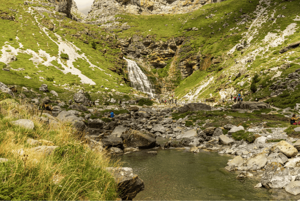 A scenic mountain waterfall cascades down a rocky cliff into a shallow stream, surrounded by green grassy slopes and scattered boulders. Hikers are visible along the trail and near the water, taking in the dramatic natural landscape under partly cloudy skies.