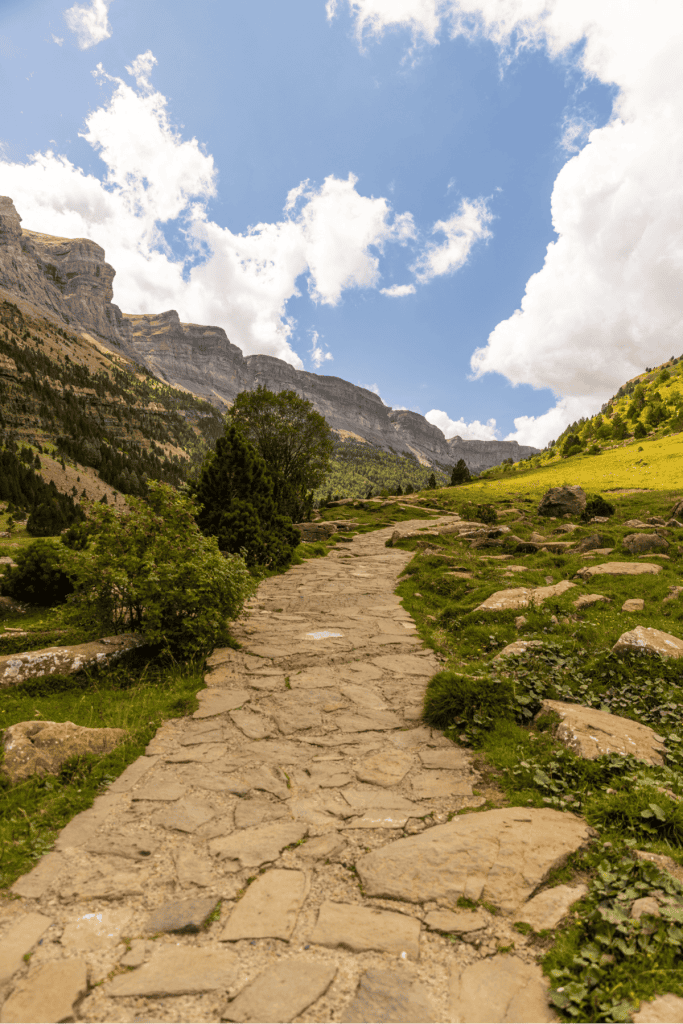 A stone path winds gently uphill through a green mountain valley, bordered by grassy slopes and scattered rocks, with dramatic cliffs rising under a bright sky filled with puffy clouds.