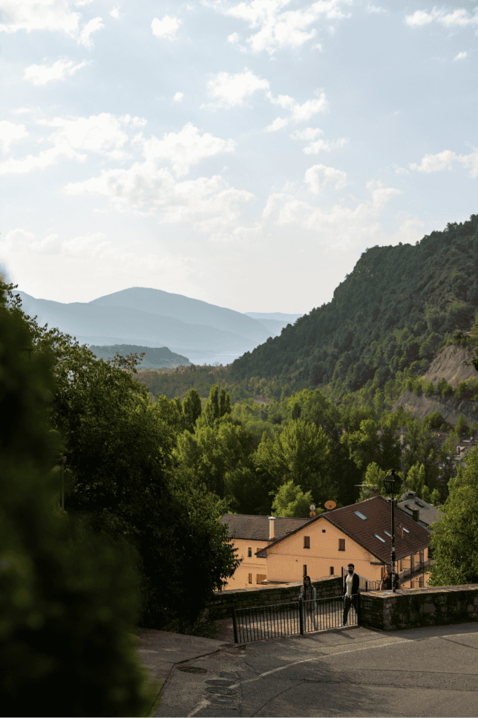 A gently curving road descends toward a cluster of peach-colored houses nestled among lush trees, with forested hills and misty mountains stretching into the hazy distance. The golden light adds a warm, tranquil feel to the quiet hillside scene.