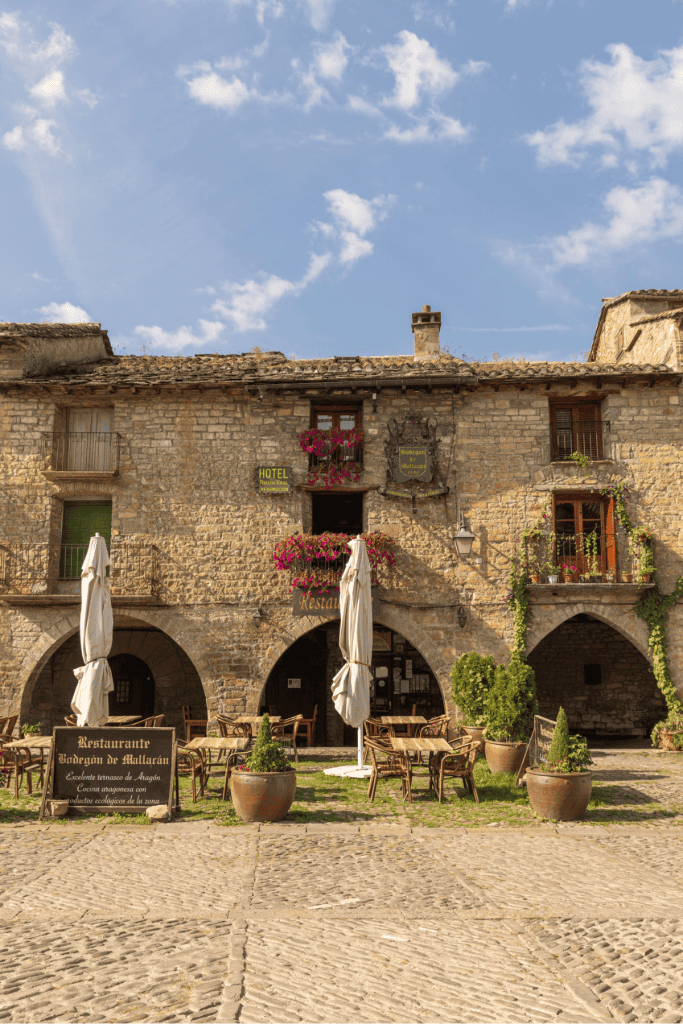A rustic stone building with arched doorways houses a charming restaurant with outdoor seating, shaded by closed white umbrellas and surrounded by potted plants. Bright pink flowers hang from the balcony above, and signs for the restaurant are mounted on the facade, adding to the cozy, historic village atmosphere.