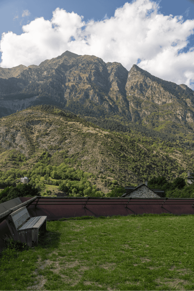 A grassy terrace with wooden benches overlooks a lush green valley and towering rocky mountains, offering a peaceful scenic viewpoint.