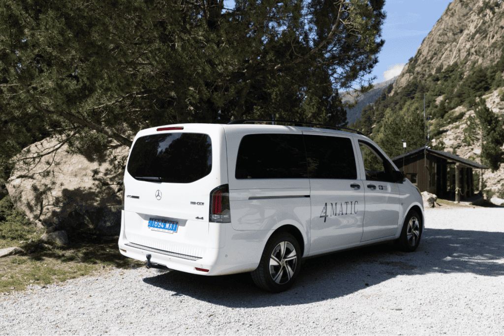 A white Mercedes-Benz van with "4MATIC" branding is parked on a gravel road surrounded by tall trees and rocky mountains. The setting suggests a scenic, mountainous location with a wooden cabin visible in the background.