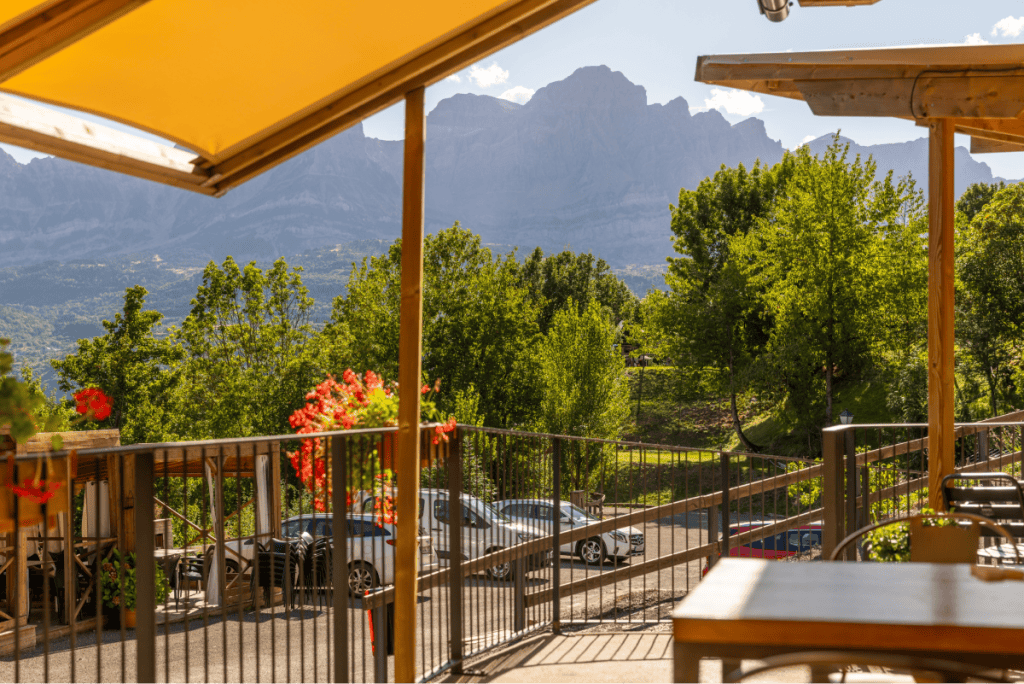 A sunny terrace with wooden tables and yellow awnings overlooks a tree-filled landscape and a parking area, with jagged mountain peaks rising dramatically in the background. Bright red flowers add a pop of color to this peaceful outdoor setting.
