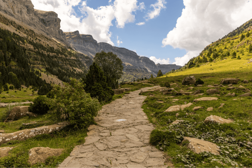 A stone-paved hiking path winds through a lush alpine valley, flanked by green slopes, scattered rocks, and forested cliffs. Towering mountains rise in the distance under a partly cloudy sky, creating a dramatic and inviting landscape for outdoor exploration.