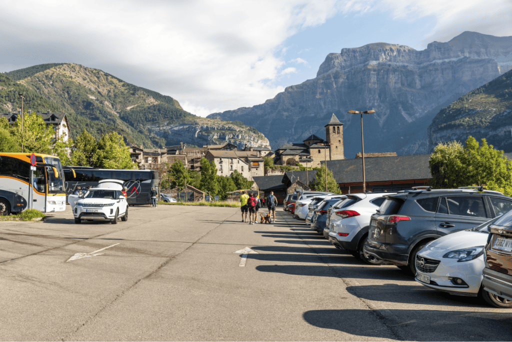 A group of hikers with backpacks and a dog walks through a sunny parking lot lined with cars, with a scenic village and dramatic mountain cliffs rising in the background. A tour bus and a historic stone church tower are visible, framed by lush green hills.