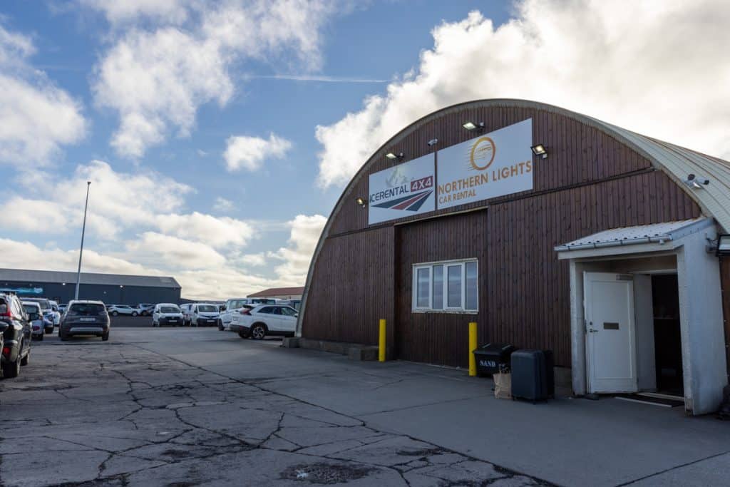 A curved wooden building houses IceRental 4x4 and Northern Lights Car Rental, with signs above a small window and entrance door. The lot is filled with parked cars under a partly cloudy sky, suggesting an airport or travel hub location.