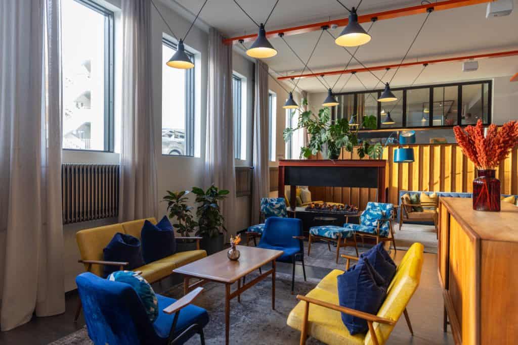 Cozy hotel lounge with mid-century modern furniture in blue and yellow tones, centered around a sleek fireplace and surrounded by large windows and leafy plants. Warm lighting and wooden accents create an inviting, retro-inspired atmosphere.