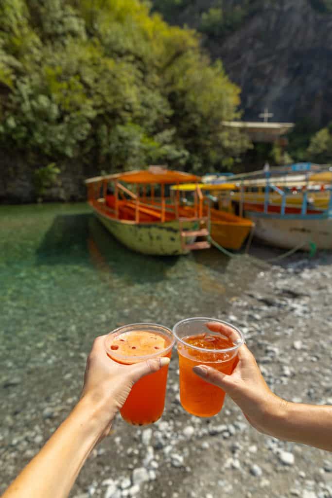 Two people clink clear plastic cups filled with a bright orange drink near the edge of a rocky riverbank. Behind them, colorful wooden boats are docked in clear turquoise water, surrounded by lush green trees and steep cliffs.