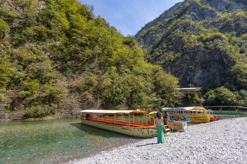 Woman standing on a rocky riverbank beside colorful wooden boats docked on the clear green waters of the Osumi River in southern Albania. Towering forested cliffs rise steeply in the background, framing the peaceful canyon setting under a bright blue sky.