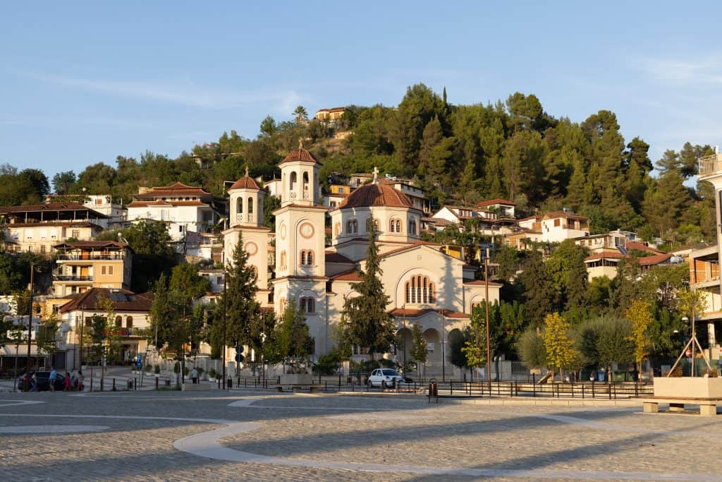 A large church with three domed towers sits in a quiet plaza at golden hour, with hillside homes and dense trees surrounding it in the background.