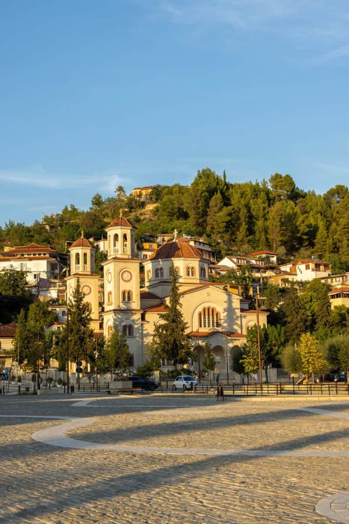 Large Orthodox church in Berat, Albania, with twin bell towers and red-tiled domes, framed by cypress trees and backed by a hillside of traditional houses and greenery. The golden hour lighting casts a warm glow across the church facade and the surrounding town square.
