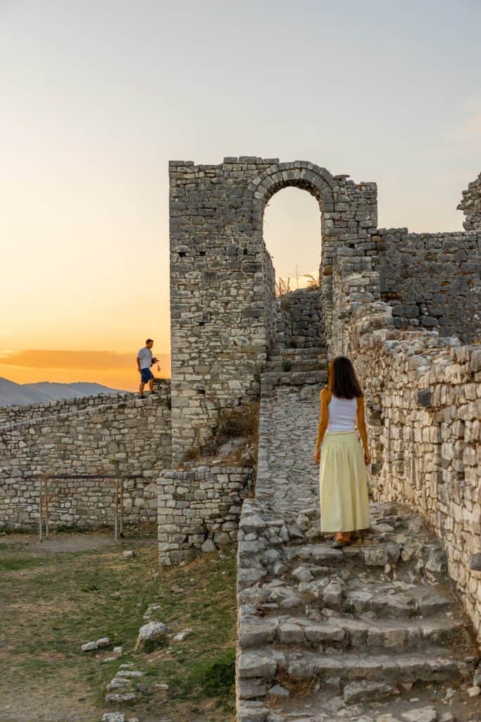 Woman in a white top and yellow skirt walks up a stone stairway inside the ancient ruins of Berat Castle at sunset. A man with a camera stands on the fortress wall nearby, silhouetted against the glowing orange sky and distant hills.
