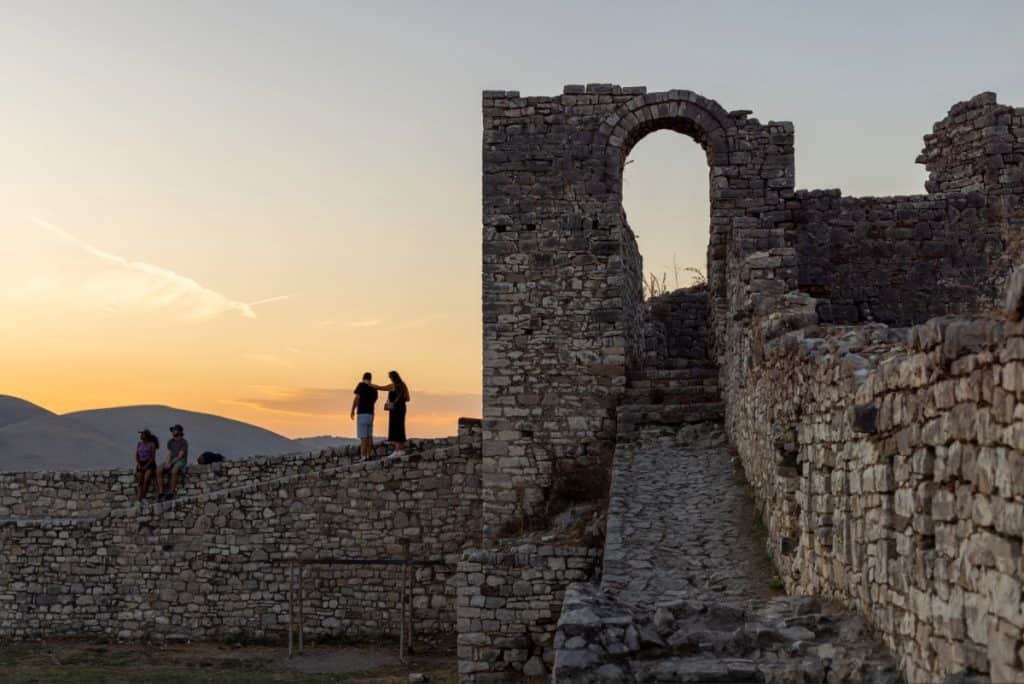 Silhouettes of visitors walking and sitting atop the ancient stone walls of Berat Castle in Albania at sunset. The warm glow of the sky highlights the weathered textures of the fortress, with a prominent arched gateway leading up a cobbled path.