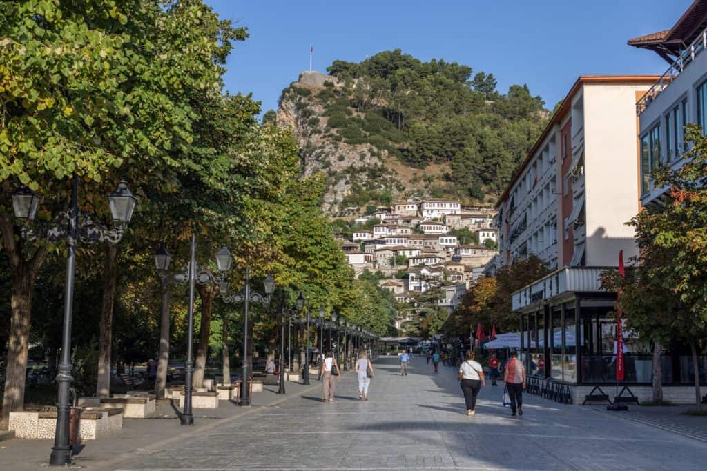 Pedestrian walkway flanked by leafy trees and vintage-style lamp posts leads toward hillside homes with white facades and red-tiled roofs. A fortress with a flag sits atop the rocky hill overlooking the town.