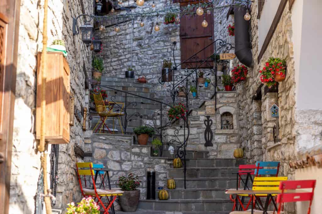 Charming stone staircase in a narrow alleyway in Berat, Albania, decorated with colorful chairs, potted plants, and hanging string lights. The cozy setting features rustic stone walls, wrought iron railings, and small pumpkins scattered along the steps for an inviting, lived-in feel.