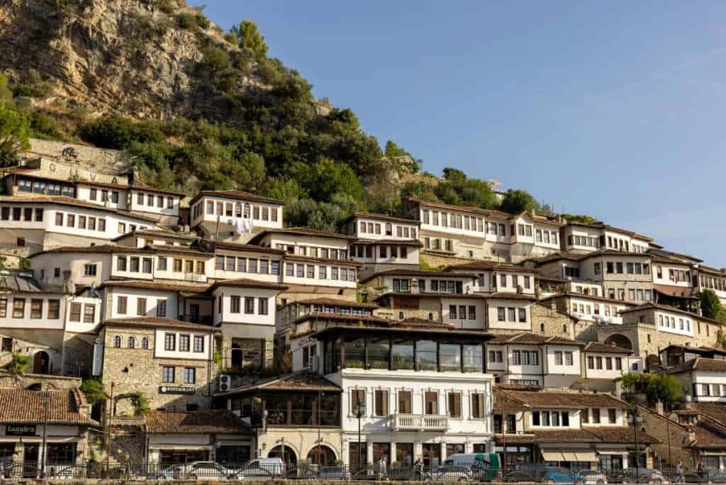 Hillside view of the historic Ottoman-era houses in Berat, Albania, known for their uniform white façades and large windows, stacked closely together on the slope beneath a rocky hill. The architecture and layout give the appearance of a “city of a thousand windows.”