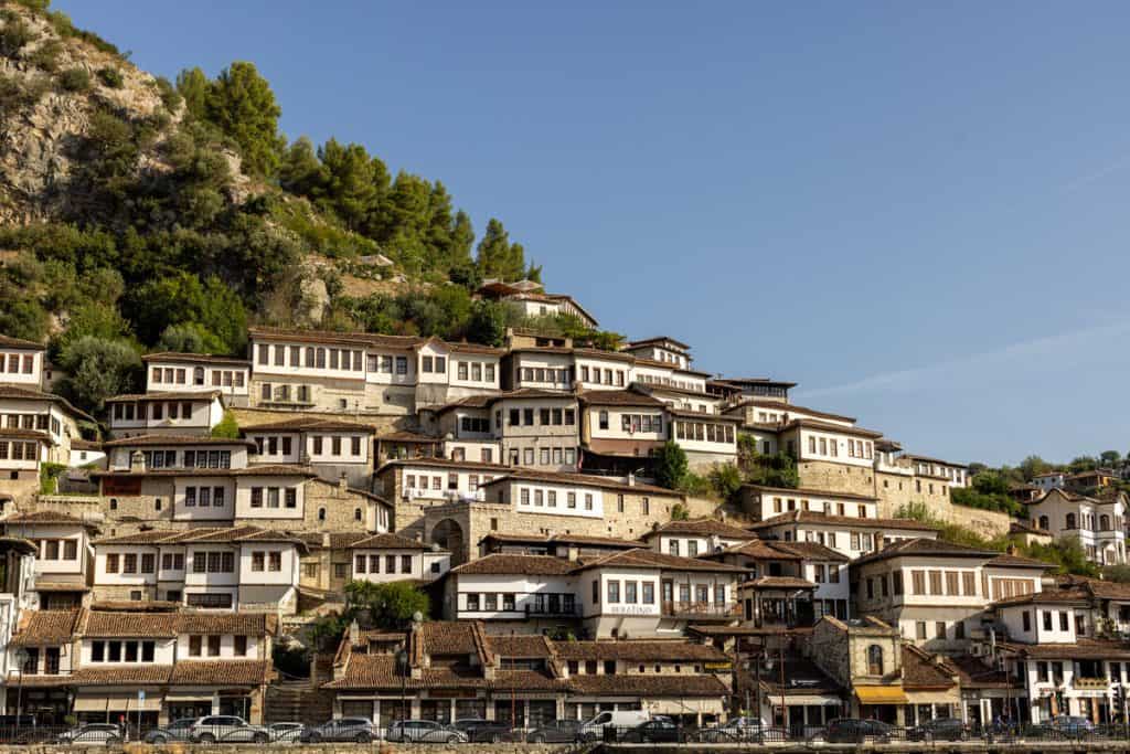 Clustered Ottoman-style white houses with brown-tiled roofs climb up the hillside in Berat, Albania, a UNESCO World Heritage site. The stacked windows and uniform architecture give the impression of “a city of a thousand windows” under clear blue skies.