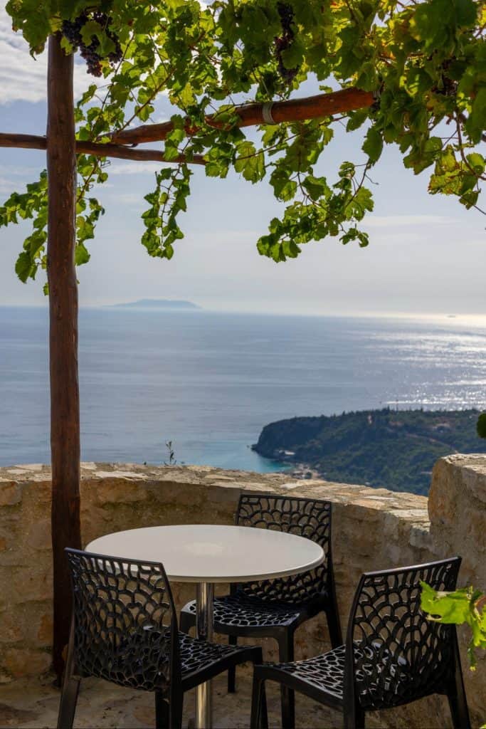 Cozy round table with four black chairs set on a stone terrace shaded by a grapevine pergola, with a scenic view of the ocean and lush green cliffs below. Sunlight reflects off the water, creating a serene, inviting atmosphere.