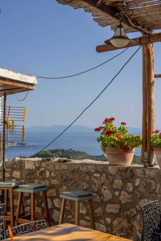 Stone terrace with wooden bar stools and potted red geraniums overlooking a calm blue sea and distant islands under a clear sky. A rustic pergola with bamboo roofing and a hanging light fixture adds charm to the coastal view.