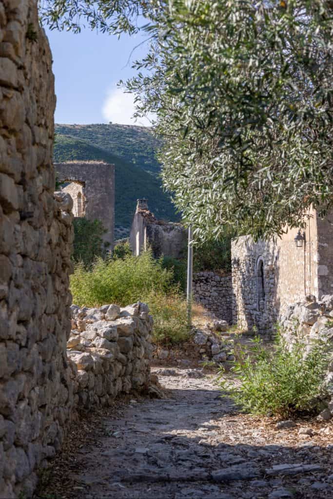 Narrow stone path lined with crumbling stone walls and overgrown vegetation in the ruins of an old village, with partial remains of buildings in the distance and green hills in the background.