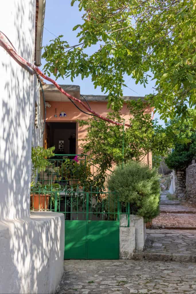Sunlit entrance to a peach-colored house in a quiet Albanian village, framed by leafy trees and a green metal gate, with potted plants and a stone path leading up to the doorway.
