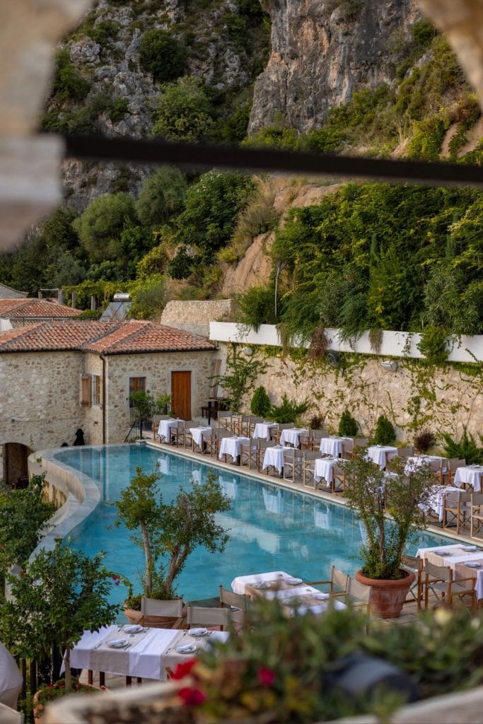 A serene pool lined with white-clothed dining tables and wooden chairs stretches alongside a stone building, set against a dramatic backdrop of lush greenery and rocky cliffs.