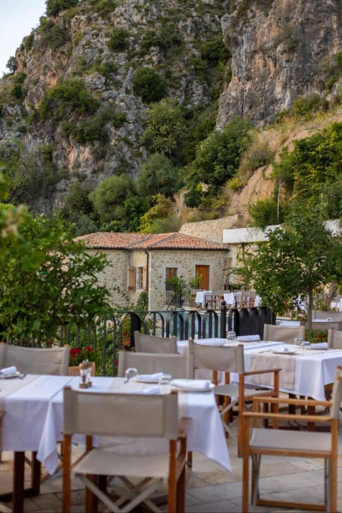 Outdoor dining tables with white linens and wooden chairs are arranged on a stone patio surrounded by greenery, with a rustic stone building and steep rocky cliffs in the background.