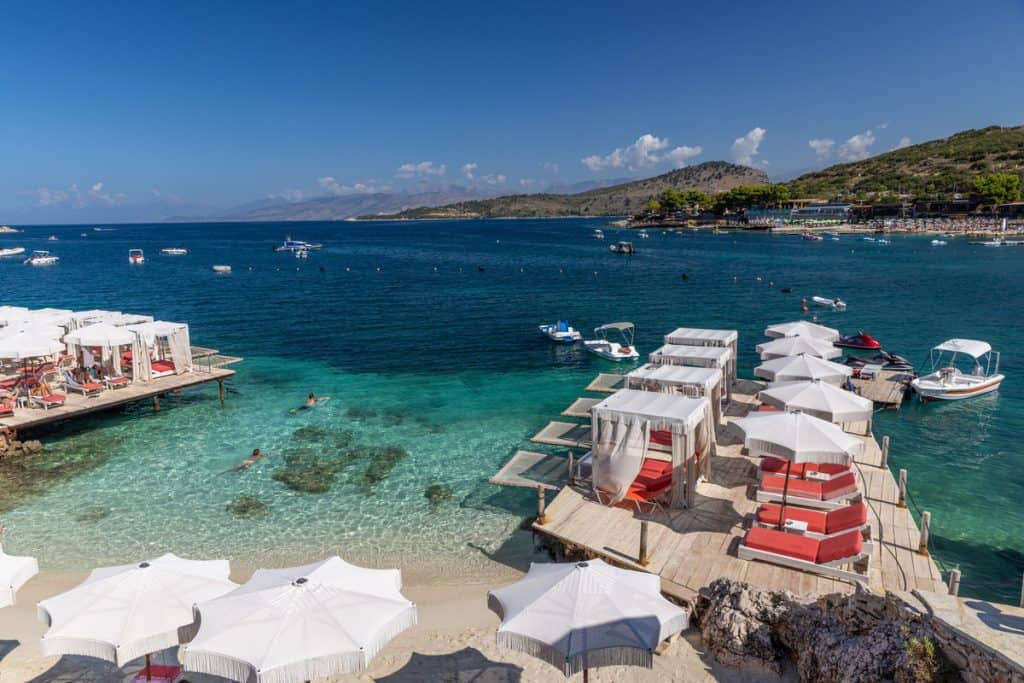 Private cabanas with red cushions and white canopies line wooden platforms over turquoise water, while swimmers and boats enjoy the bay under a bright blue sky.