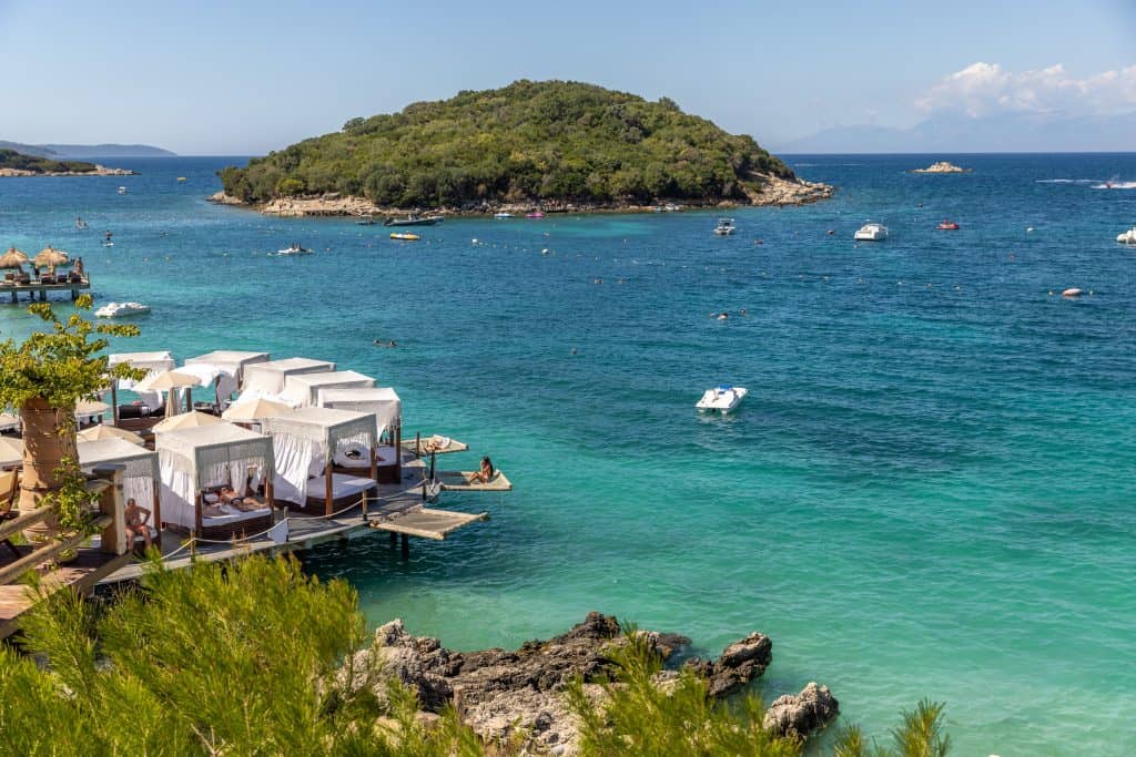 Luxury cabanas line a wooden deck over clear turquoise water, with people sunbathing beside the shore. In the background, small boats float near a lush green island under a clear blue sky.