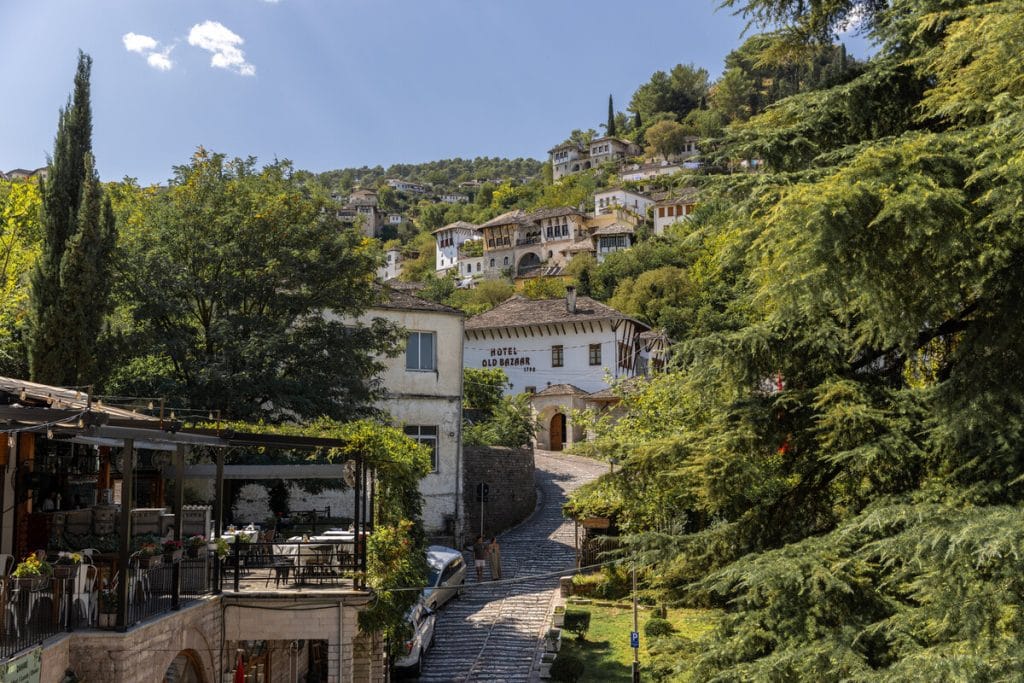 View of a cobblestone path leading uphill through the lush greenery of Gjirokastër, Albania, with traditional Ottoman-style white stone houses scattered on the hillside. The "Hotel Old Bazaar" sign is visible on one of the buildings near the center, and an open-air restaurant with empty tables sits in the foreground.