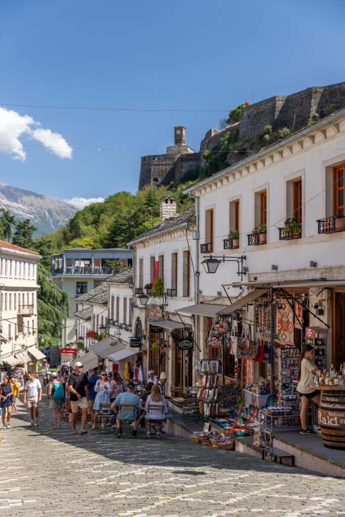Busy cobblestone street in the historic center of Gjirokastër, Albania, lined with Ottoman-style white buildings, local shops selling handmade goods, and a view of the Gjirokastër Castle on the hill above.