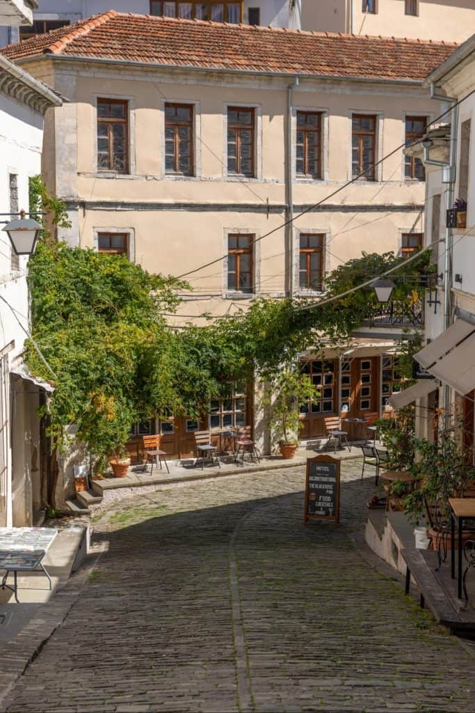 Quiet cobblestone alley in Gjirokastër, Albania, shaded by leafy vines and lined with small cafés and rustic buildings with wooden windows and terracotta rooftops.