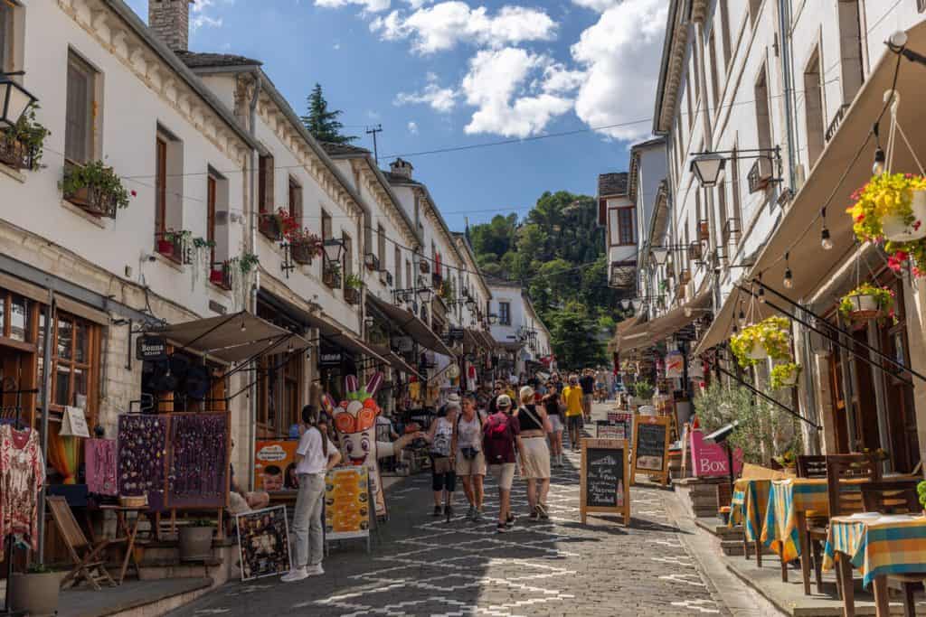 Bustling pedestrian street in Gjirokastër’s Old Bazaar, lined with white stone buildings, outdoor cafés, and souvenir shops. Tourists walk along the cobblestones, surrounded by colorful textiles, potted flowers, and hanging signs.
