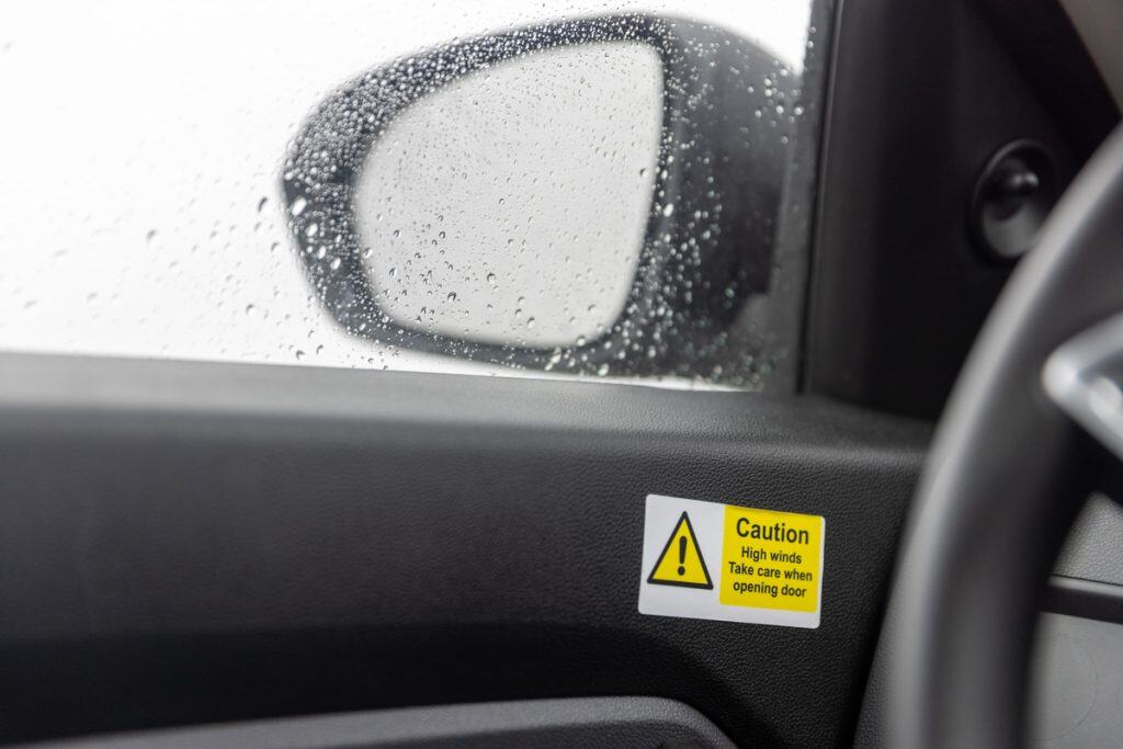 Interior view of a car door with a rain-covered window and side mirror visible, featuring a yellow caution sticker that reads "Caution. High winds. Take care when opening door." The warning highlights safety precautions in windy weather.