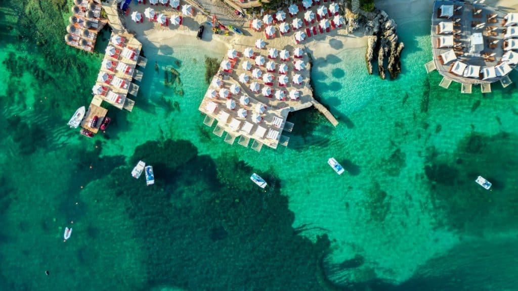 Aerial view of a beach resort in Ksamil, Albania, with clear turquoise waters, white umbrellas and lounge chairs arranged on wooden decks and sandy shores, and several small boats floating nearby.