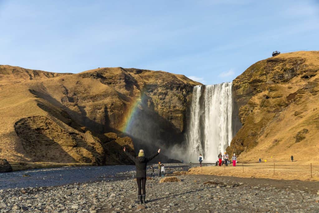 A person with arms raised stands on a rocky riverbank facing Skógafoss waterfall in Iceland, with a vivid rainbow forming in the mist to the left of the cascading water. Golden hills surround the falls, and several tourists explore the scenic area under a clear blue sky.