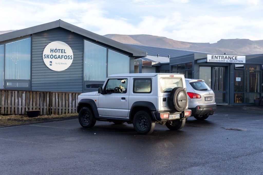 Two vehicles are parked outside Hótel Skógafoss, a modern building with metal siding and large windows, located near the mountains in Iceland. A sign above the entrance reads “Restaurant & Reception,” indicating guest amenities within the hotel.