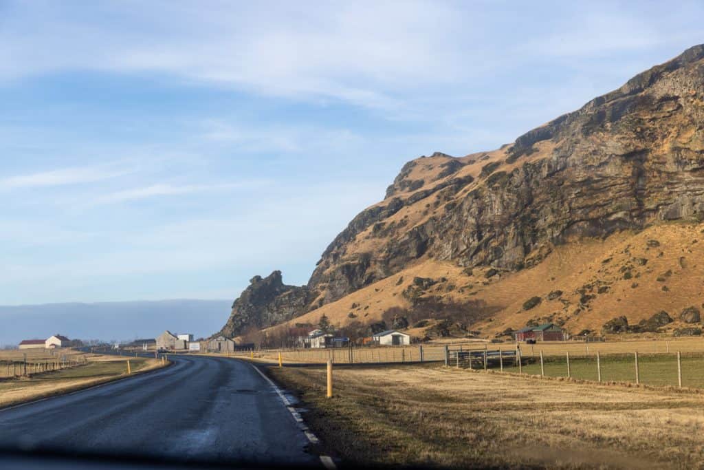 A winding road leads toward a cluster of rural buildings at the base of a rugged, golden-brown cliff under a blue sky. The scene captures a quiet countryside landscape with dramatic natural rock formations.