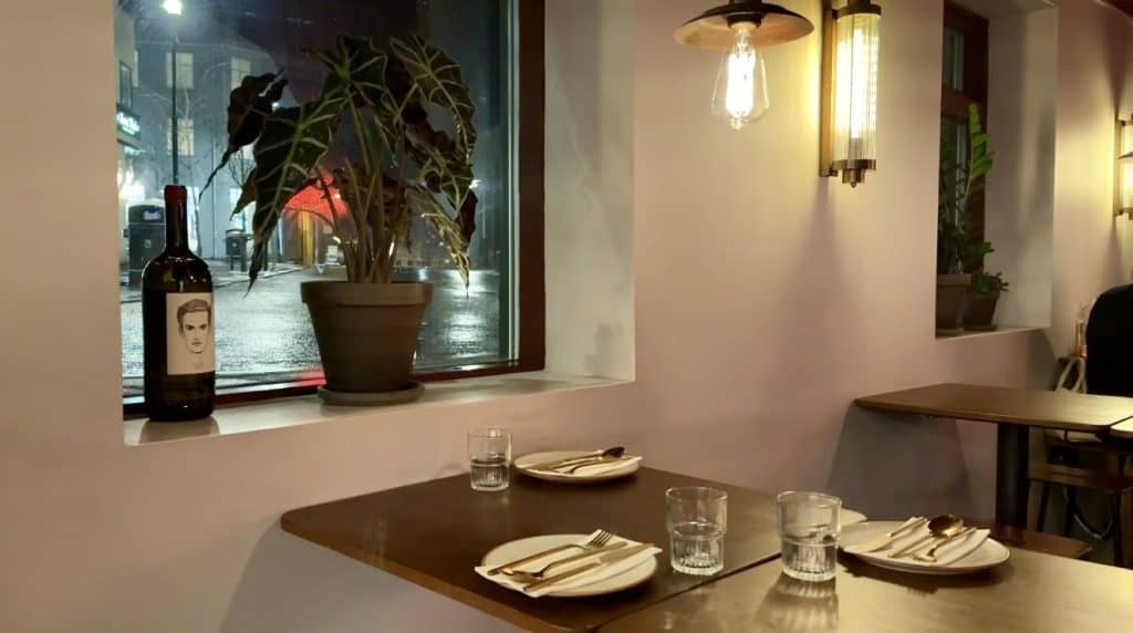 Minimalist restaurant interior featuring a wooden table set for two, warm lighting, and a potted plant beside a window overlooking a rainy city street. A wine bottle with a sketched portrait on the label sits on the windowsill.