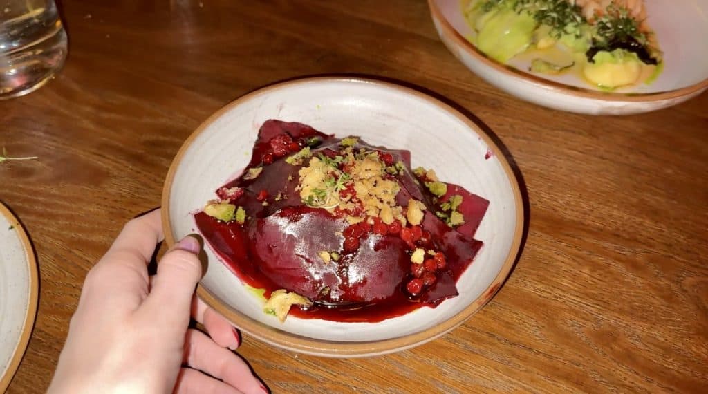 A plate of beetroot ravioli topped with crushed nuts, fresh herbs, and a glossy red sauce is held by a hand over a wooden table. Another small plate of food is partially visible in the background.
