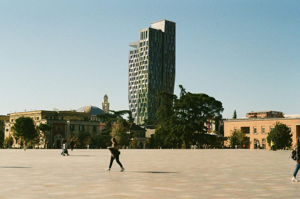 Wide view of Skanderbeg Square in Tirana, Albania, with people walking across the open plaza. In the background, the modern green-glass Plaza Tirana hotel tower contrasts with older, historic buildings and tall trees.