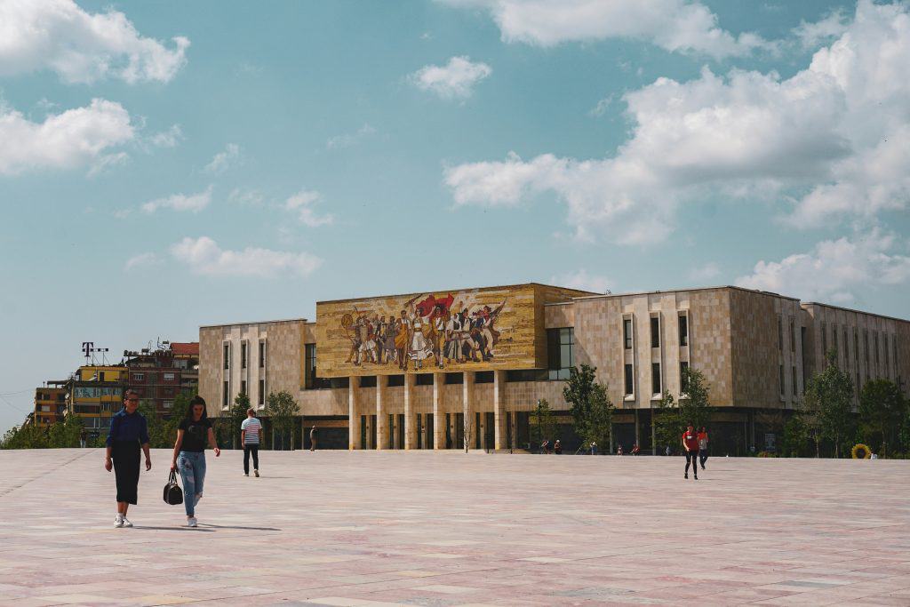 Skanderbeg Square in Tirana, Albania, featuring the National History Museum with its iconic mosaic mural depicting Albanian historical figures. A few people are scattered across the spacious plaza under a partly cloudy sky.